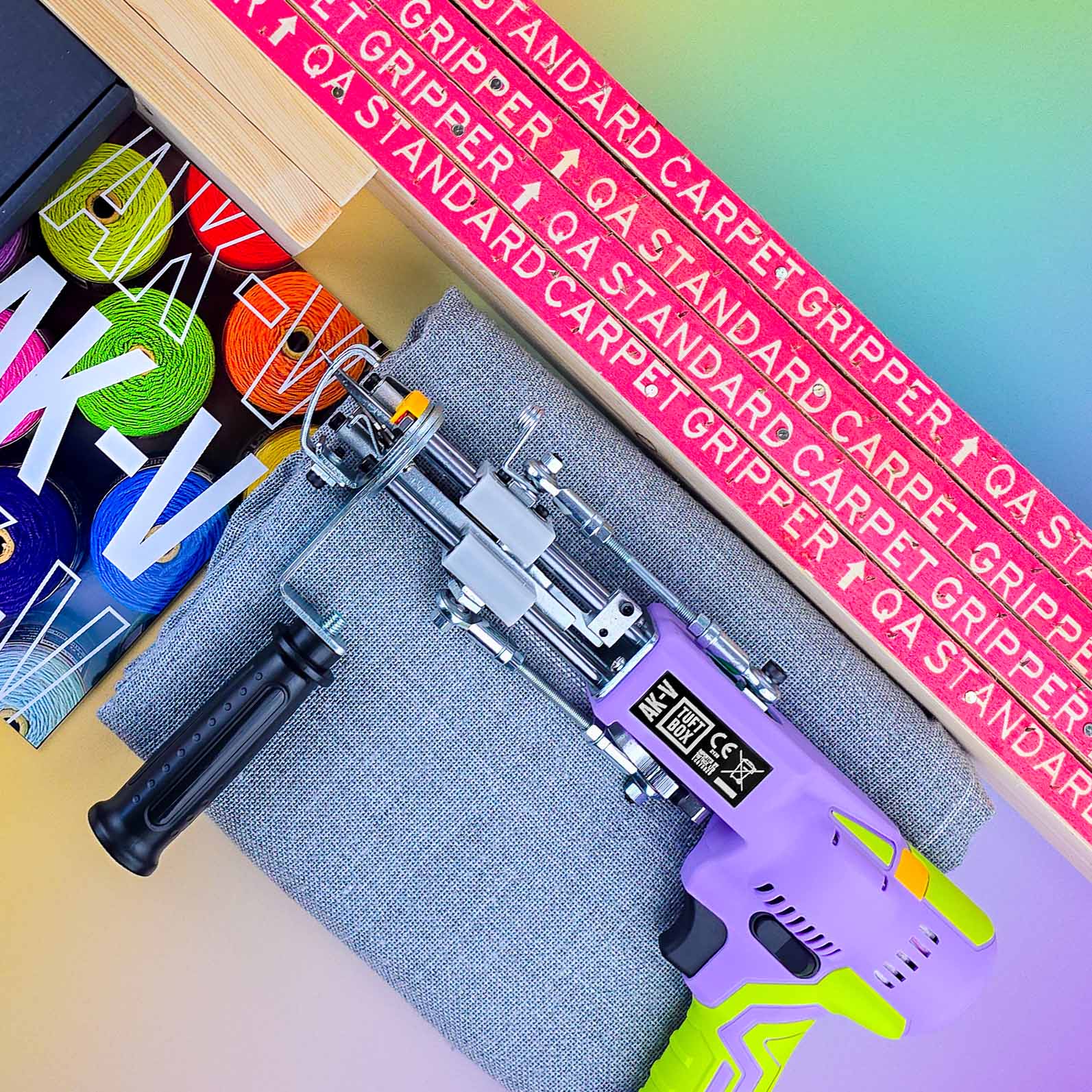 Purple AK-V 5 rug tuft machine laying on tufting fabric next to a wooden rug tufting frame on a soft pastel coloured gradient background