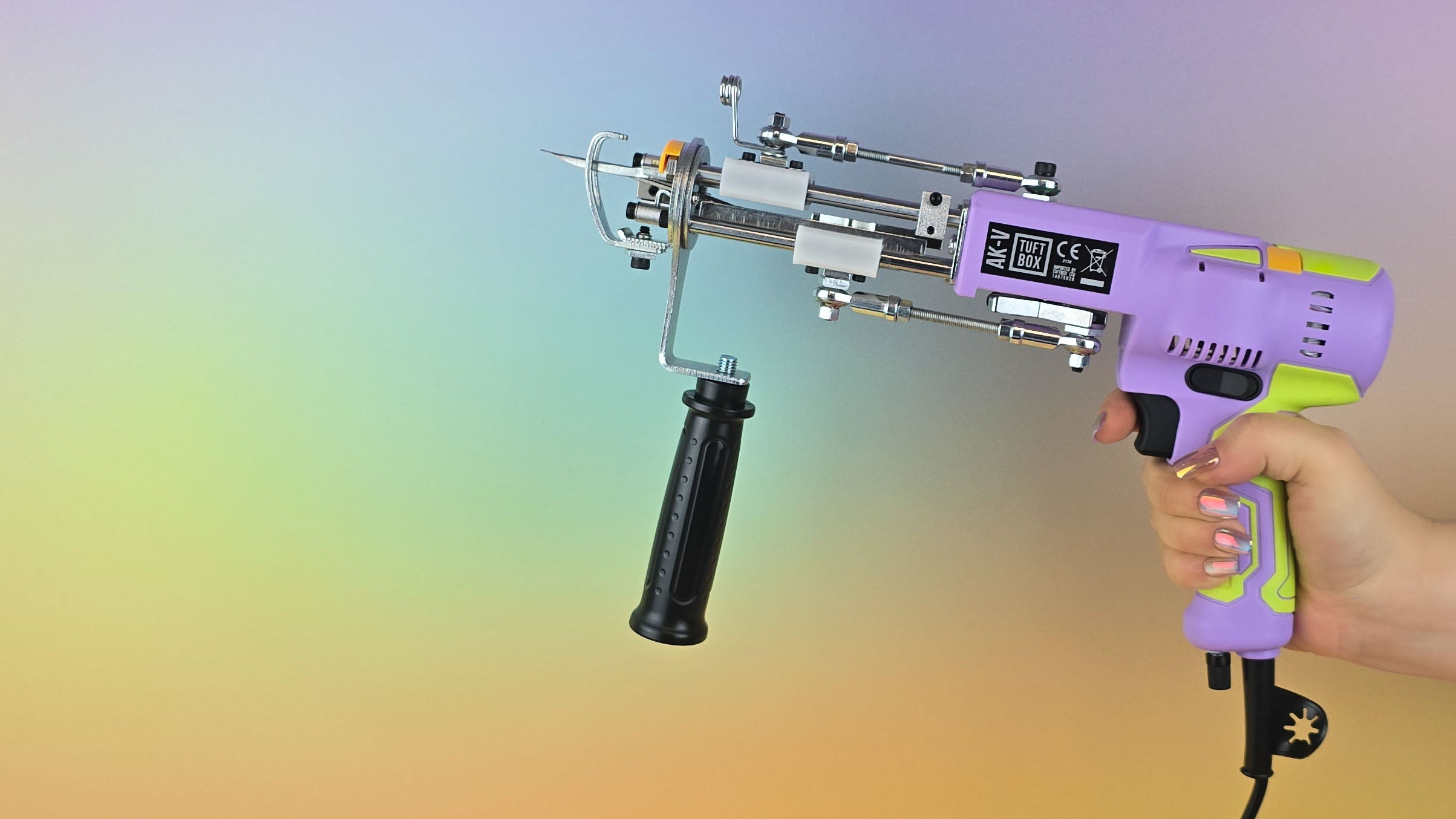 Hand holding a purple and green AK-V rug tufting machine power tool against a soft rainbow gradient background