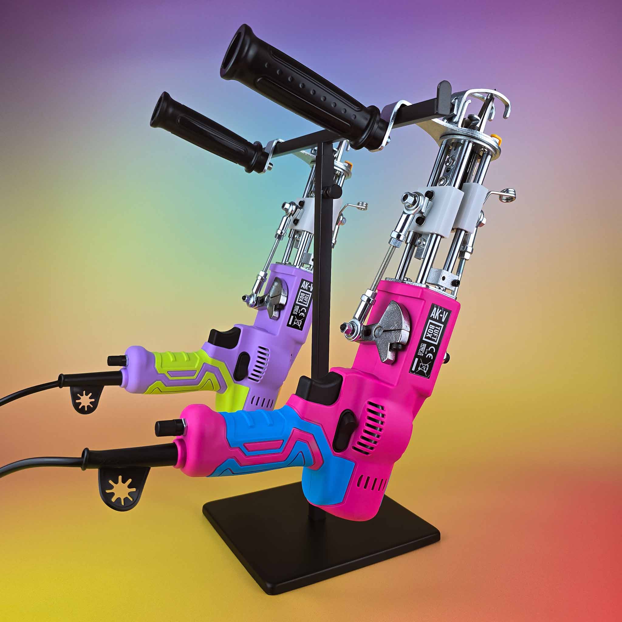 Pink and purple colourful AKV AK-5 rug tufting guns balanced on a black stand with gradient rainbow backdrop