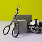 Black Crane Embroidery Scissors leaning on grey blocks with a black/white pom pom
